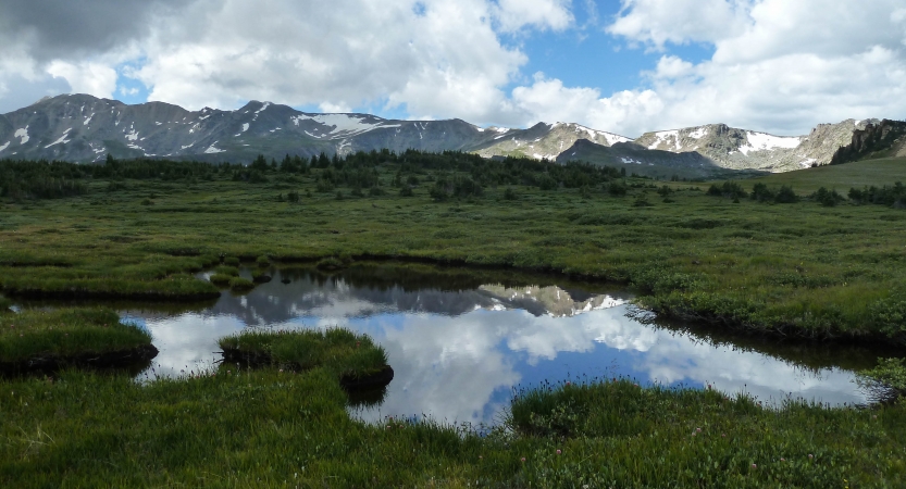 a rocky and snowy mountain ridge is reflected in the still waters of a pond